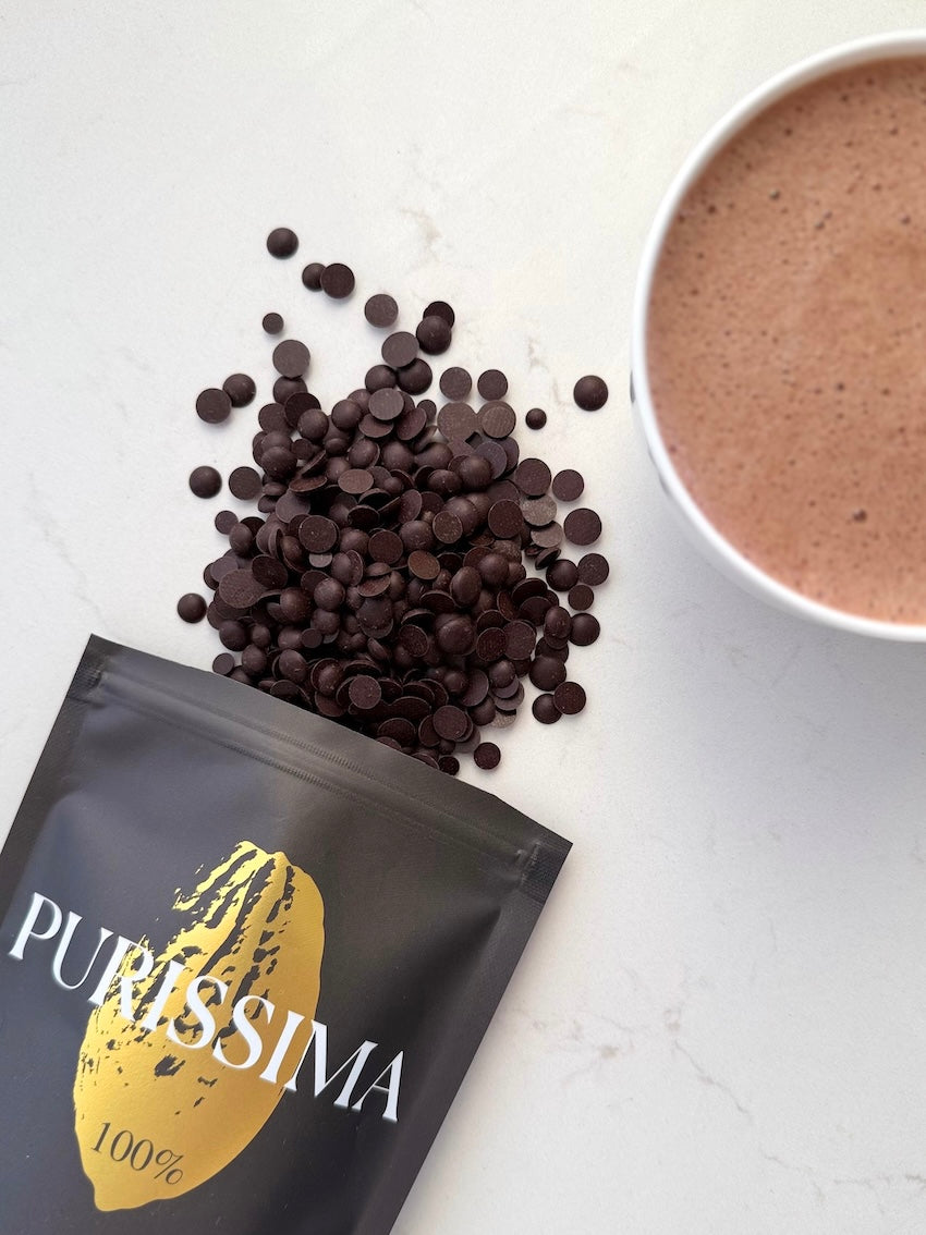 Verpackung der Sorte Purissima, dunkle Schokoladendrops und eine Tasse mit aufgeschäumter purer Trinkschokolade 100% Kakao.