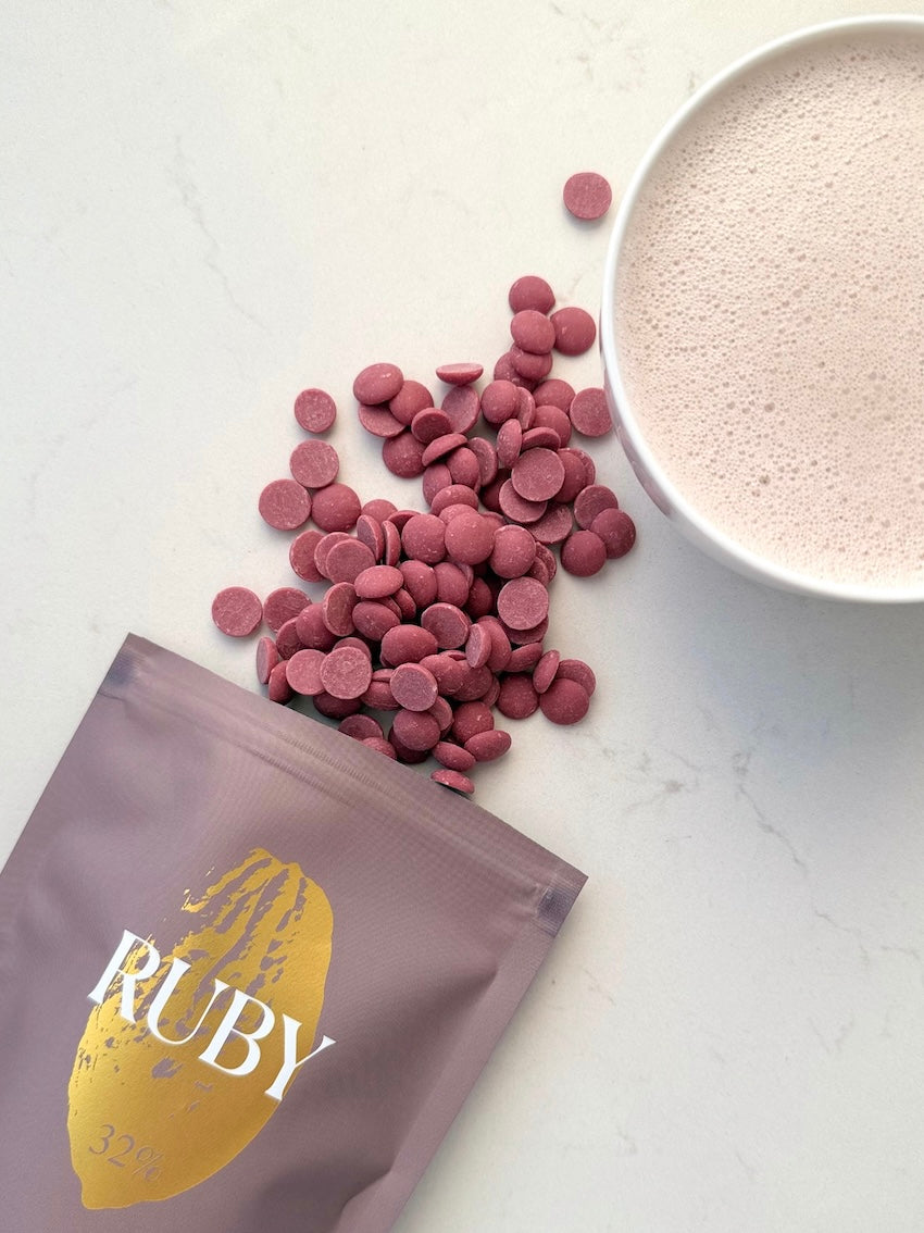 Verpackung der Sorte Ruby, rosa Schokoladendrops und eine Tasse mit aufgeschäumter Ruby Trinkschokolade.