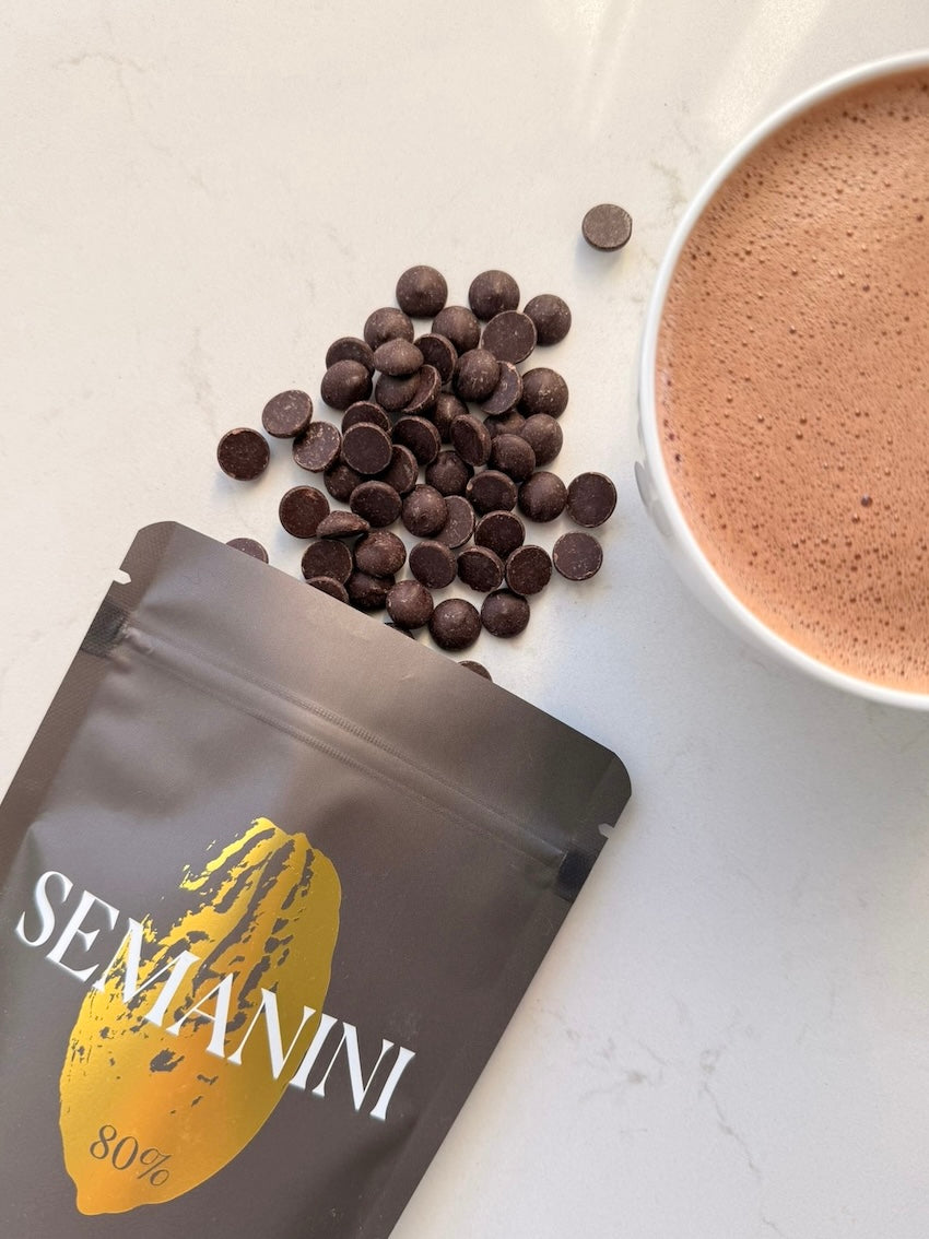 Verpackung der Sorte Semanini, dunkle Schokoladendrops und eine Tasse mit aufgeschäumter dunkler Trinkschokolade mit 80 % Kakao.